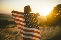 a woman holding an American flag standing in a field at sunset 