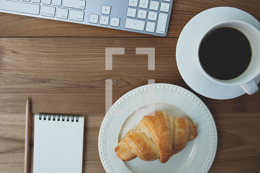 croissant on a plate, notepad and pencil, computer keyboard, and coffee mug on a table 