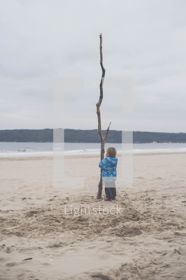 a toddler holding a large stick on a beach in a winter coat 
