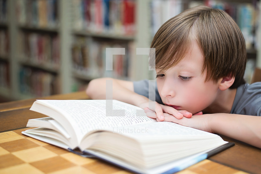 Boy reading a book on a table in a library.