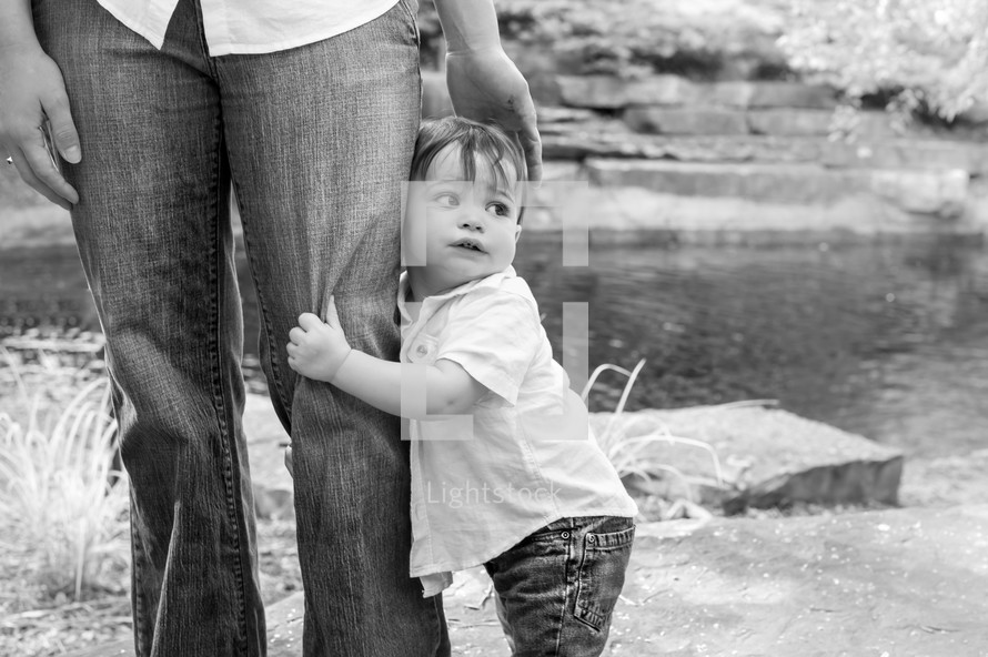 Boy clinging to his father's leg while standing outside.
