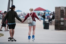 an adorable couple holding hands on a beach boardwalk while they roller blade and skateboard 