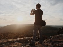 man on a mountaintop taking a picture of a sunset 