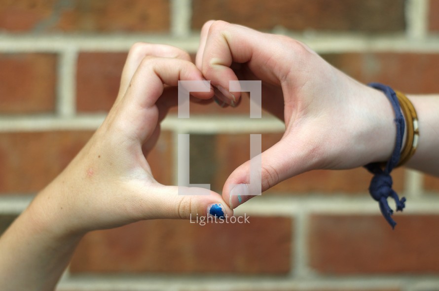 Heart hands in front of a brick wall.