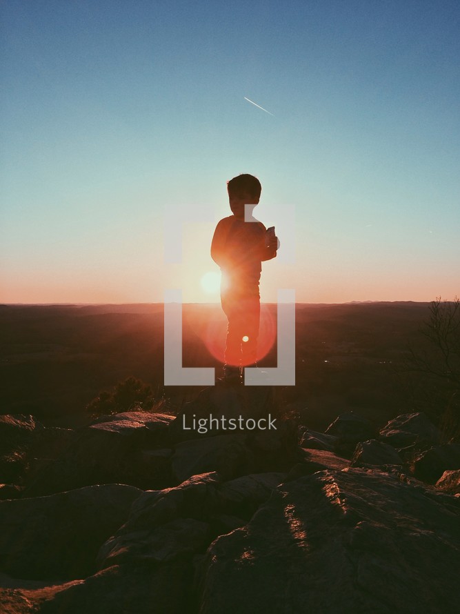 A silhouette of a young boy standing at the top of a mountain under intense sunlight. 