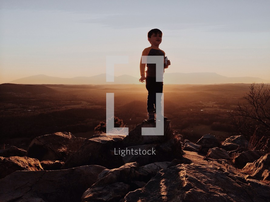 Silhouette of a child standing atop a rock in front of a mountain range at dusk.