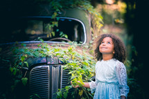 a little girl standing in front of a vintage car covering in ivy 