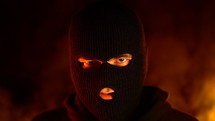 Portrait Of Young Man In Black Balaclava Against Backdrop Of Blazing Night Fire