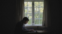 Man reading Bible at desk and taking notes on laptop in front of a window with natural light