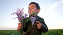 Portrait of cute little boy in festive costume with butterfly and bouquet on open area field background. Child, handsome son with flowers, holiday concept. Sunset time. High quality 4k footage