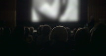 Back view of people watching movie in the cinema. Defocused screen with film lightening viewers in dark hall