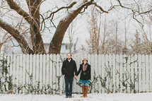 a couple standing in front of a fence in the snow holding each others hands 