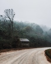 fog over trees and cabin on the side of a dirt road 