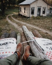 feet on a pile of logs and cabin near a dirt road 