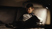 a little boy sitting on a couch reading a Bible 