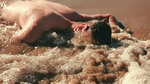 a man face down on a beach as water washes over him 