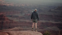Male hiker walks to the edge of a cliff and admires stunning canyon views during sunset