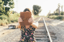 A girl walking down an abandon railroad track and holding her bible.