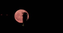 Red full moon rising from behind a large oil refinery