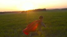 Boy dressed with a Superman cape running in a field, looking into the sunset.