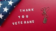 "Thank You Veterans" spelled out with white letters on a red background with an American Flag and dog tag.