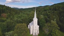 Flying by white church tower with steel cross on top in green forest nature, christian religion temple
