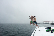 Three teens jumping from a snow covered bank into the ocean.