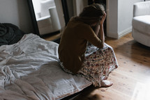 a woman with her hands on her head sitting at the edge of a bed 