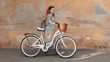 Beautiful young woman wearing long dress walking the old european street near retro white bicycle with basket. Slow Motion.