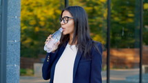 Young businesswoman near office building drinks mineral water from bottle. Beautiful girl in eyeglasses drinking clean water. Healthy lifestyle concept.