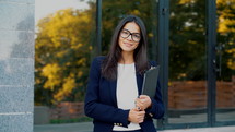 Portrait of young beautiful businesswoman looking at camera with smile outdoors, professional female manager wearing glasses and suit, holds documents folder. Student girl concept.