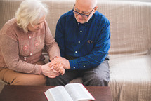 elderly couple holding hands praying and reading a Bible 