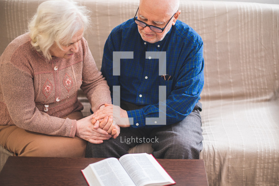 elderly couple holding hands praying and reading a Bible 