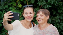 Pretty granny and her granddaughter doing selfie outdoor, looking at camera and laughing to smartphone camera. Technology, memory, family concept