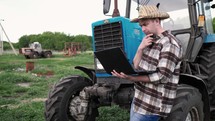 Farmer with laptop stood by tractor. Young farmer engineer with laptop standing on sun. This is a close up of a farmer using a computer on his tractor.