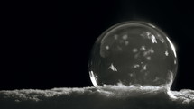 Ice crystals forming on freezing bubble sitting on snow in front of black background.
