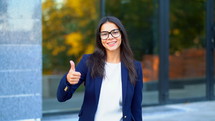 Winner. Success. Brunette young girl in business wear on office building background smiles to camera and gives thumbs up. Happy woman showing gesture of approval