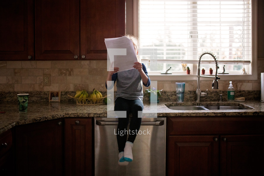 a girl sitting on a countertop in a kitchen reading papers 