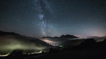 Stars with milky way galaxy moving over mountains above foggy valley in starry sky night time lapse
