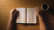 An overhead shot of a man flipping through his bible at a table with coffee