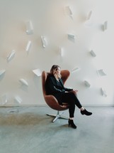 A woman sitting in a leather chair in a bare room with white sheets of paper on a white wall.