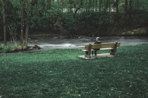 a woman sitting on a bench next to a fast flowing stream 