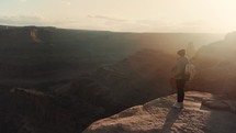 Female hiker walks to the edge of a cliff and admires stunning canyon views during sunset