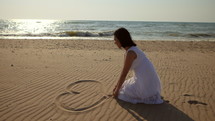 Woman in a white dress drawing a heart in the sand of a beach.