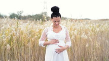 Young pretty pregnant woman posing on nature background. Expectant mother waiting for a baby. She holds and strokes her tummy with love. Girl have unusual hairstyle - braids