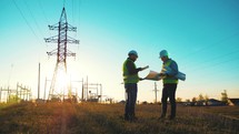Teamwork industry a concept. Two engineers electric experts in protective helmets discussing the construction of a power line. Silhouette Of Engineers Standing On Field With Electricity Towers.