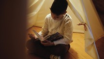 a boy child reading a children's Bible in a tent in his house 