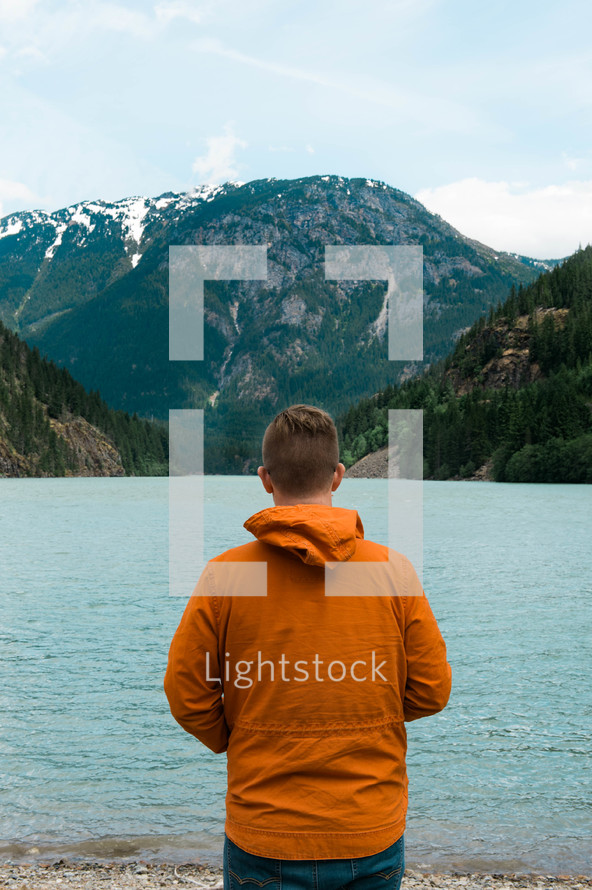 a man in an orange jacket standing by a mountain lake 