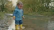 Cute baby in raincoat and rubber boots has fun by river, throws pebbles and laughs. Funny boy learns and explores natural world. Family, kids, love concept