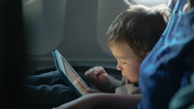 Little boy traveling in an airplane sitting in his seat playing with a tablet computer watched by a parent